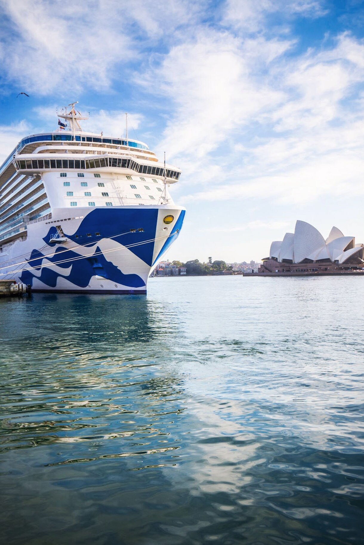 A large cruise ship docked in a calm harbor, with the iconic Sydney Opera House visible in the background under a bright, partly cloudy sky. The ship, featuring a blue and white wave design, reflects on the water's surface.