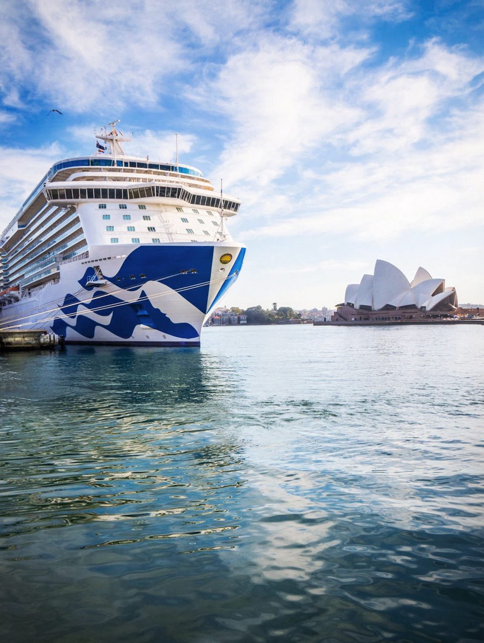 A large cruise ship docked in a calm harbor, with the iconic Sydney Opera House visible in the background under a bright, partly cloudy sky. The ship, featuring a blue and white wave design, reflects on the water's surface.