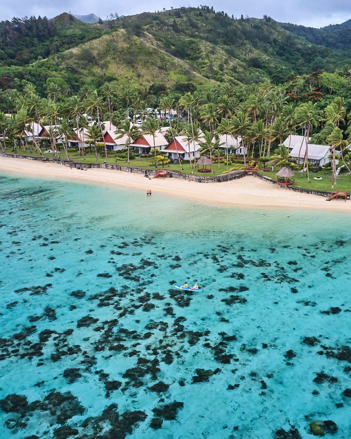 Aerial view of turquoise lagoon and sandy beach lined with palm trees and beachfront villas in Fiji, with kayakers gliding over the shallow coral waters.