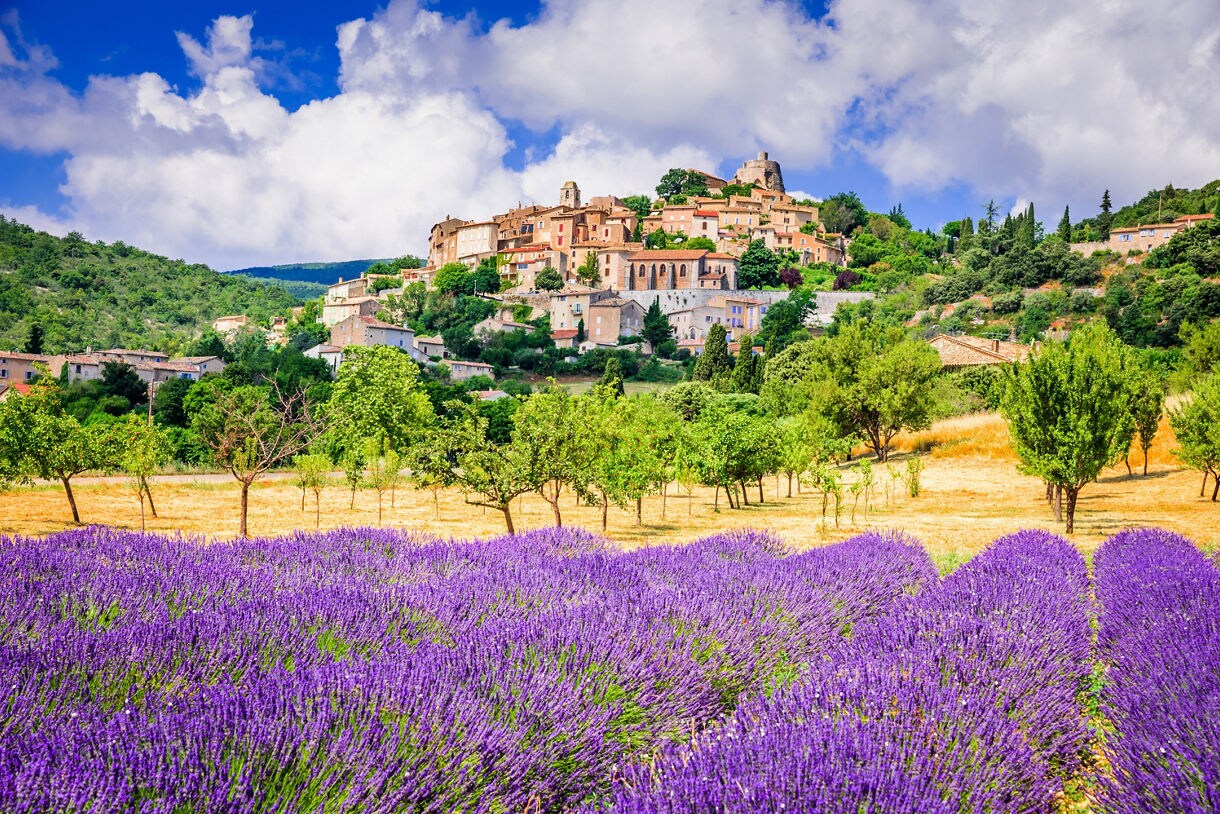 Purple lavender fields leading up to a small stone hilltop village in Provence, surrounded by green trees and rugged countryside under a bright blue sky.