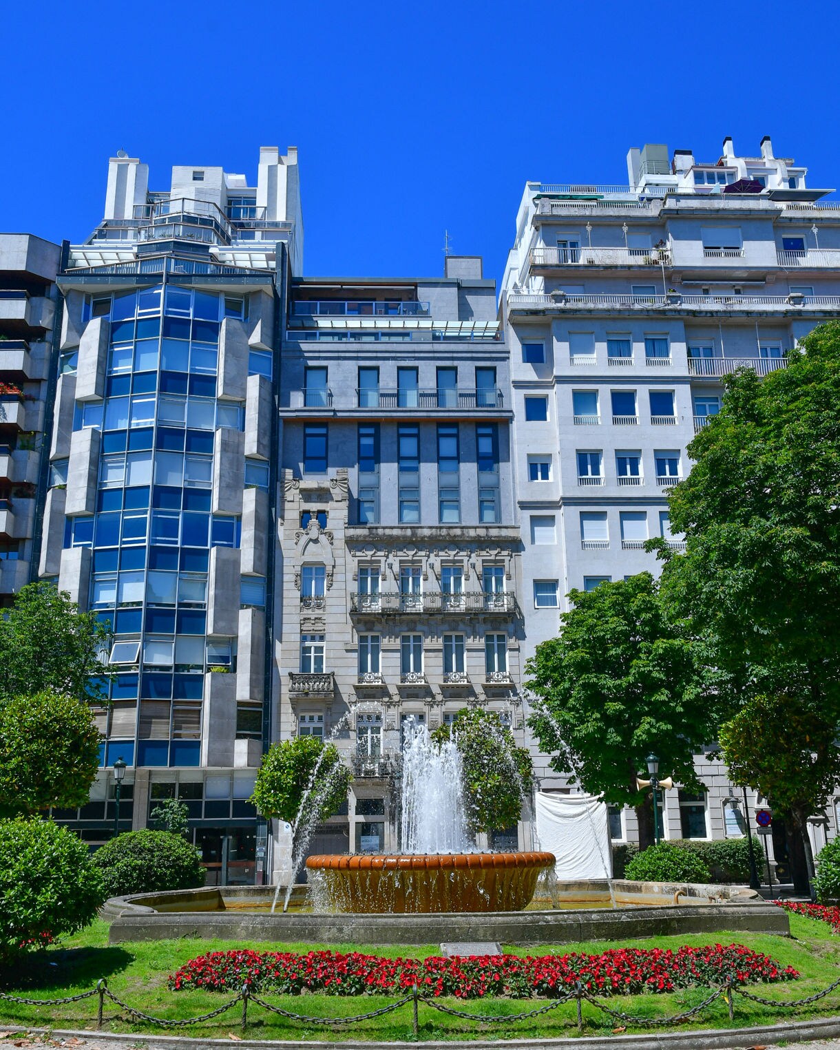 A city park with a round fountain spraying water, surrounded by grass, flowerbeds and tall buildings under a clear blue sky.