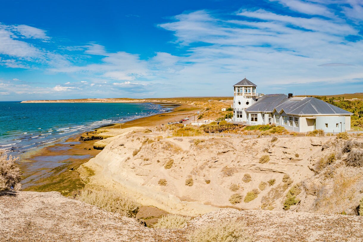 Panoramic view of Puerto Madryn’s coastline with turquoise water, sandy cliffs, dry shrubs and a white building with a tower overlooking the sea under a bright blue sky.