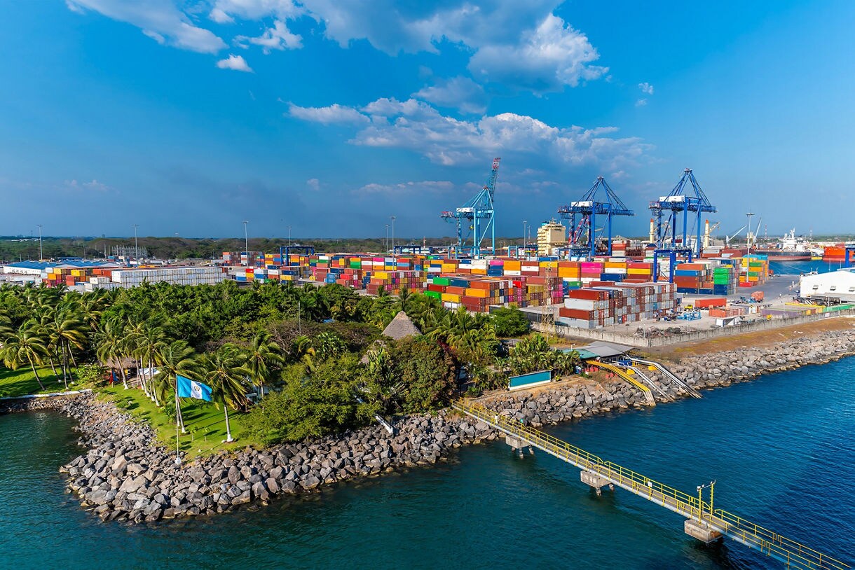 Aerial view of Puerto Quetzal in Guatemala showing a busy port with cranes, stacked shipping containers, palm trees and turquoise coastal water under a bright blue sky.