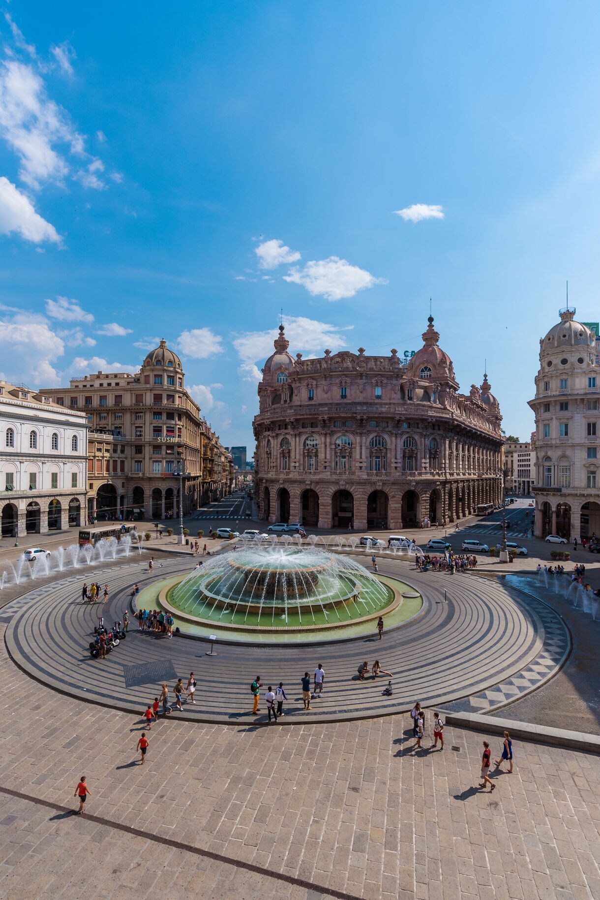 Wide view of Piazza De Ferrari in Genoa with its circular fountain surrounded by ornate historic buildings under a clear blue sky.