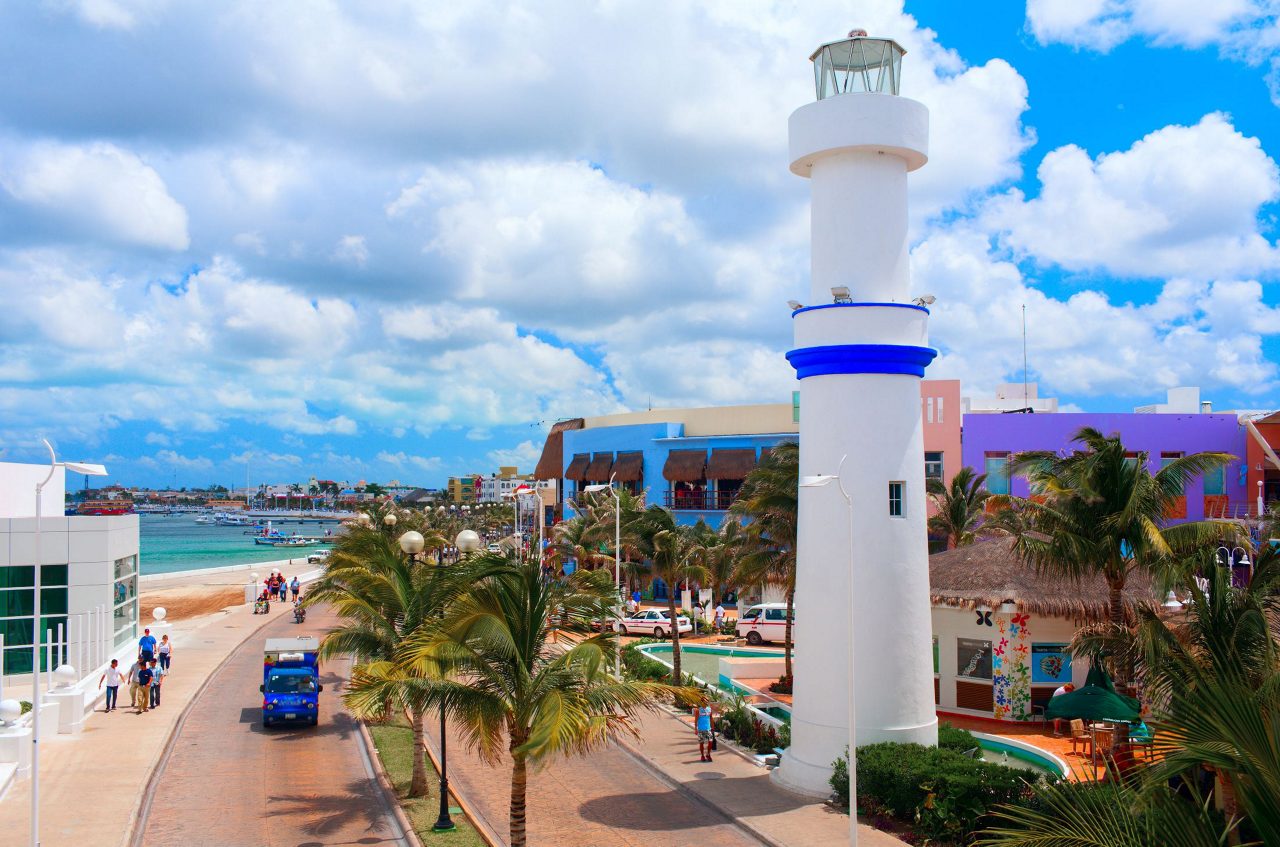 White lighthouse with blue stripe standing prominently in San Miguel de Cozumel with colorful buildings, palm trees, and Caribbean beach visible in background.