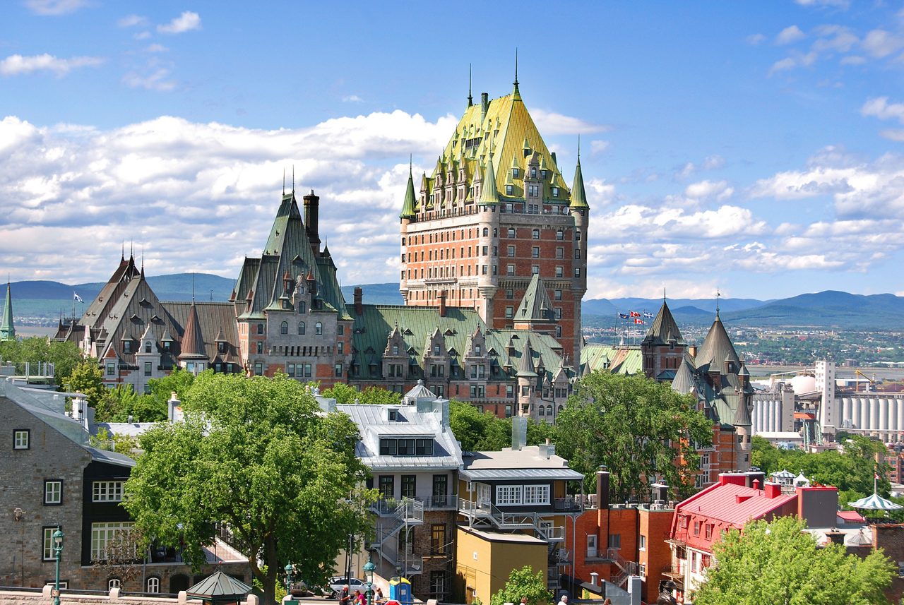 A panoramic view of Quebec City, featuring the iconic Château Frontenac with its grand architecture and green rooftops, set against a backdrop of mountains and a vibrant cityscape.