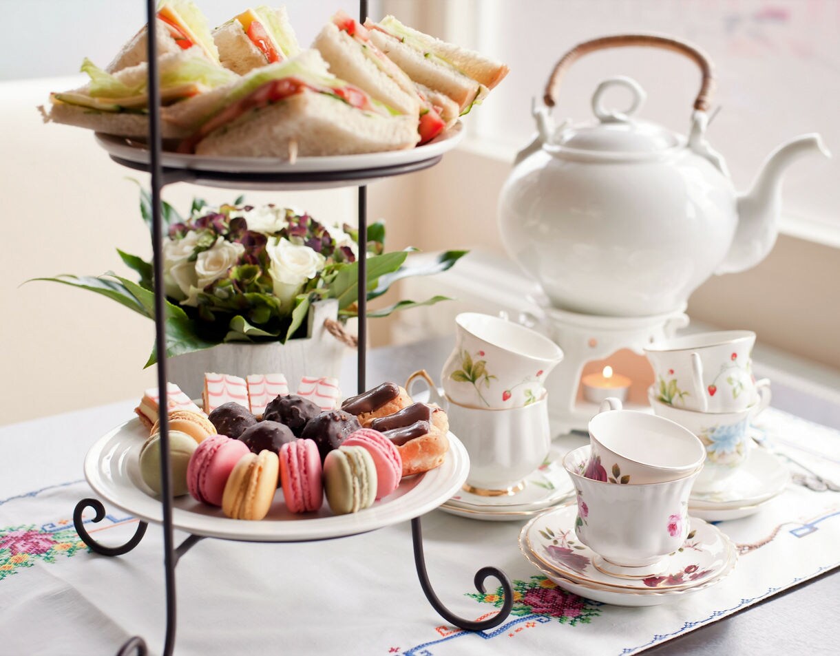 Tiered tray with macarons, pastries and sandwiches beside fine china teacups and teapot.