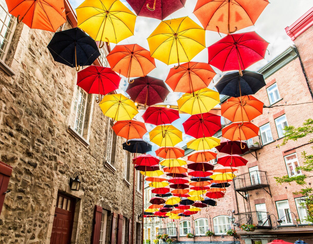 Overhead installation of red, yellow and orange umbrellas between stone buildings in Old Québec.