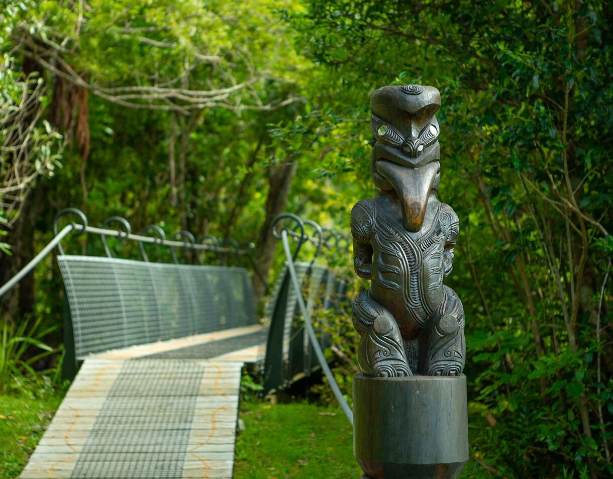 A carved Māori wooden figure stands beside a narrow boardwalk and suspension-style bridge surrounded by lush green forest.