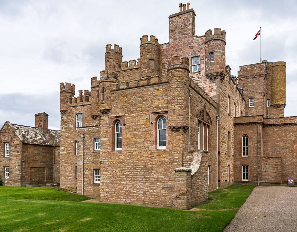 Stone-built Castle of Mey in Caithness, Scotland, featuring battlements, towers and arched windows with a Union Jack flag flying above.