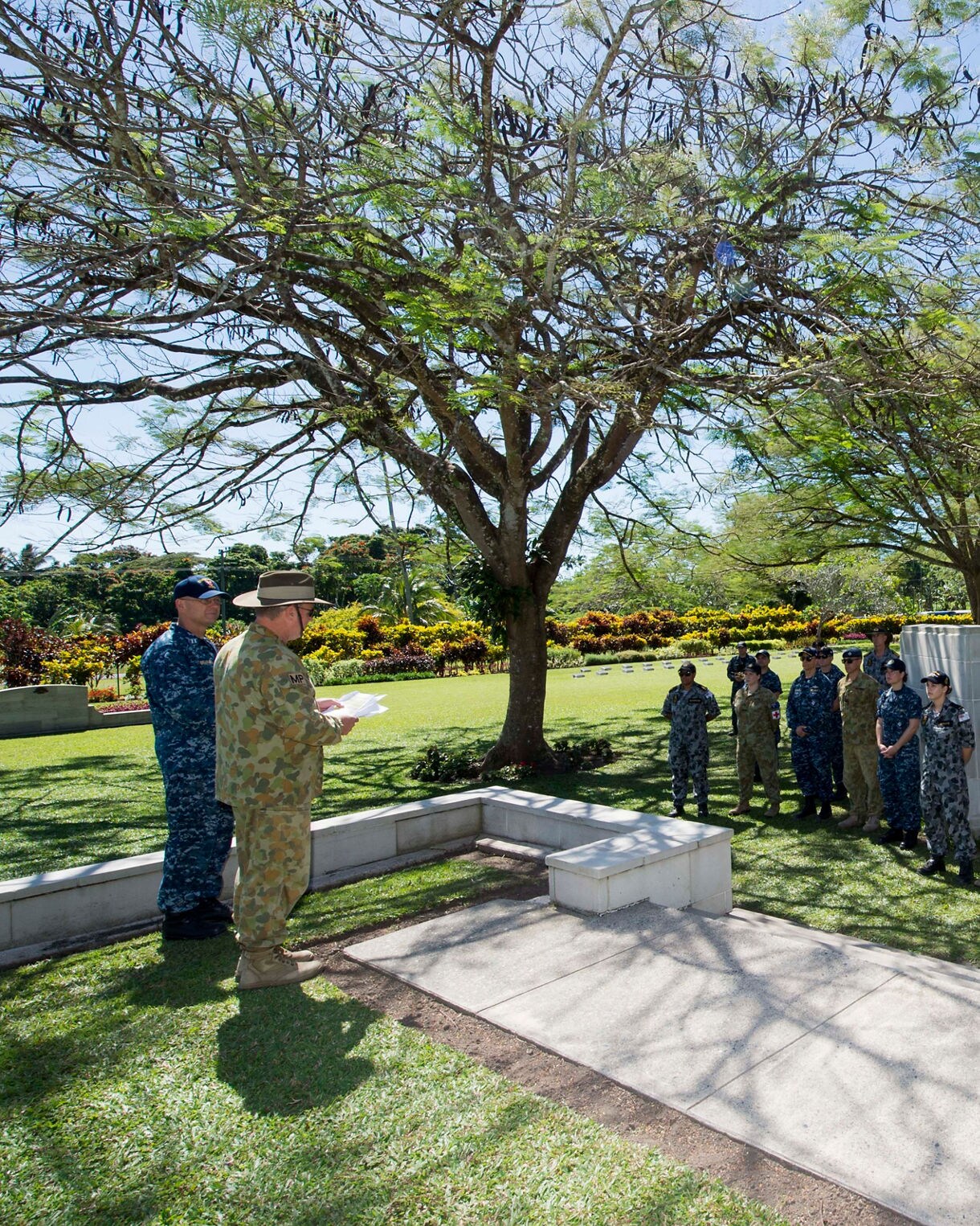 A group of uniformed military personnel stand in formation at the Bita Paka War Cemetery in Rabaul, holding wreaths during a memorial ceremony. Tall trees cast dappled shade across the lawn and stone structures frame the scene.