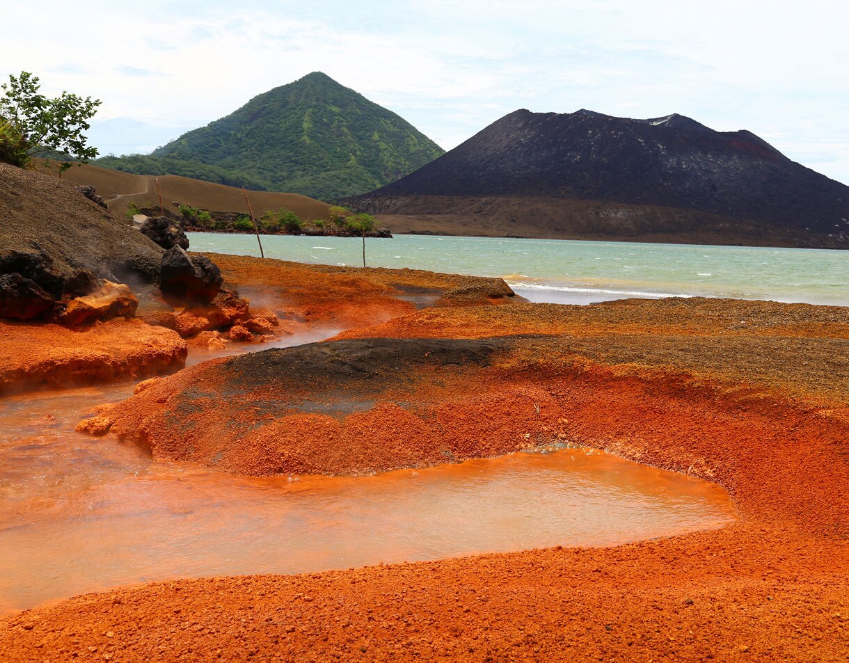 A geothermal beach in Rabaul with bright orange mineral sand, steaming hot pools and a volcanic mountain range in the background next to a green hillside.