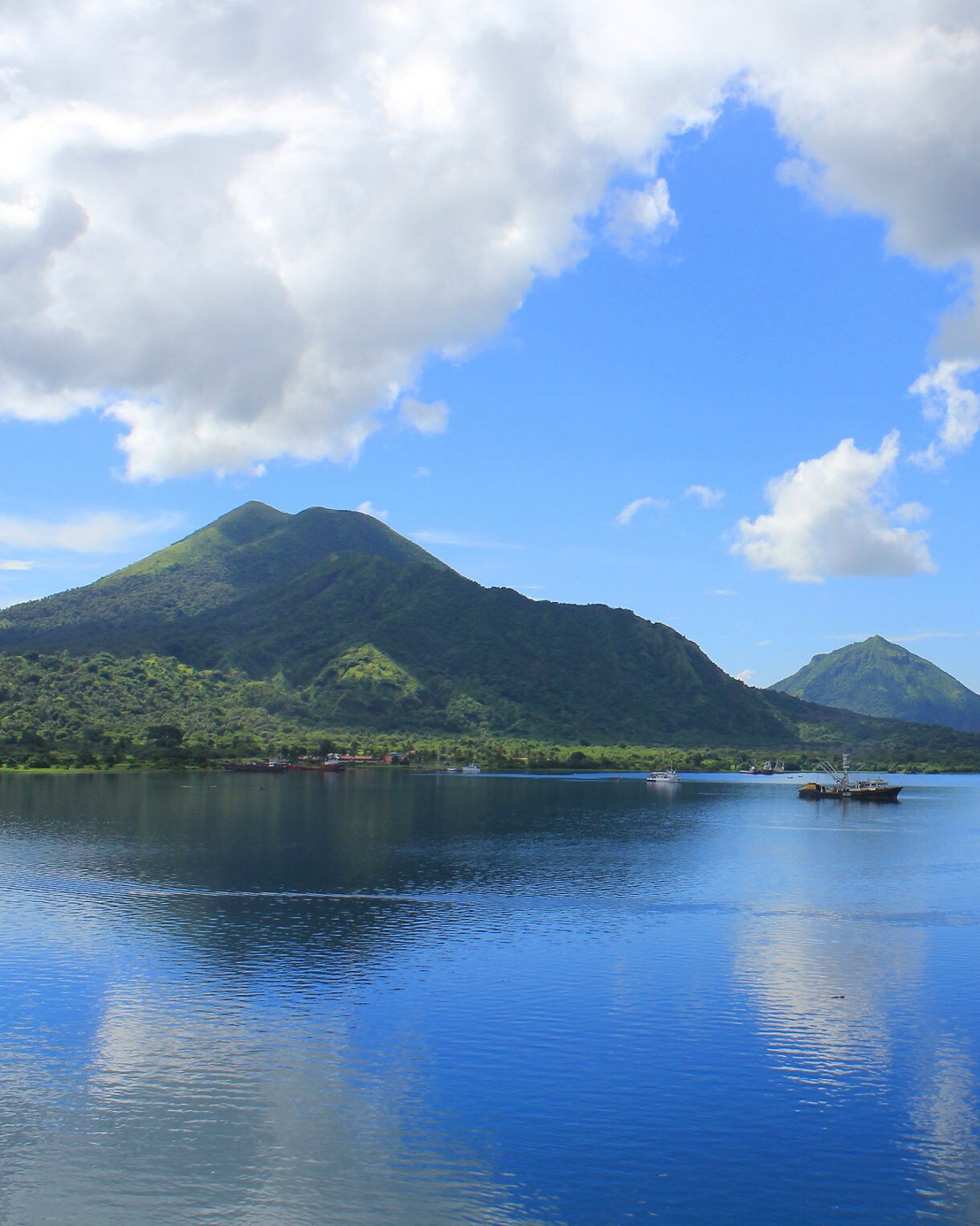 Wide view of Rabaul’s coastline featuring bright blue water, green volcanic mountains and scattered clouds reflecting on the surface.