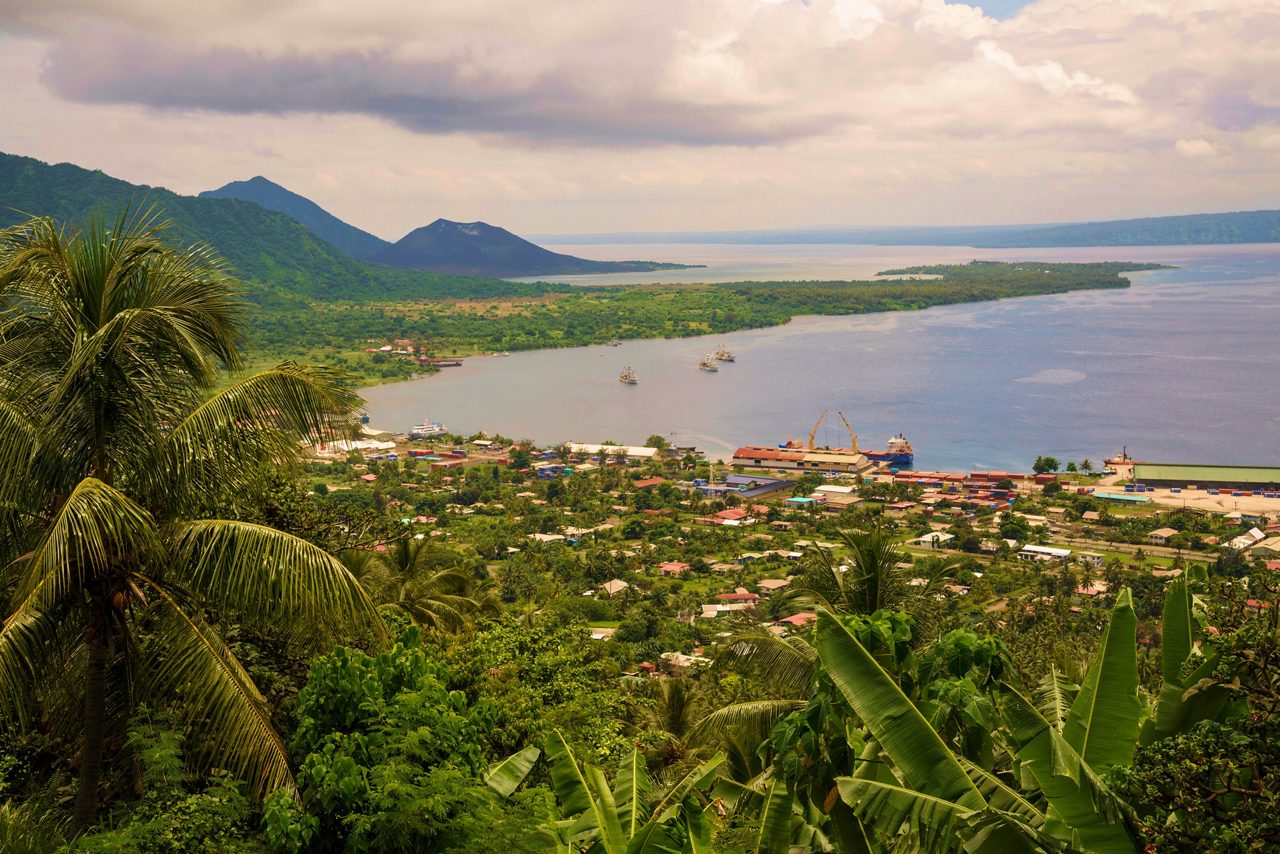 A panoramic view of a tropical coastline with dense green jungle in the foreground, a small coastal town and port below, calm blue water stretching into the distance, and volcanic mountains rising under a cloudy sky.