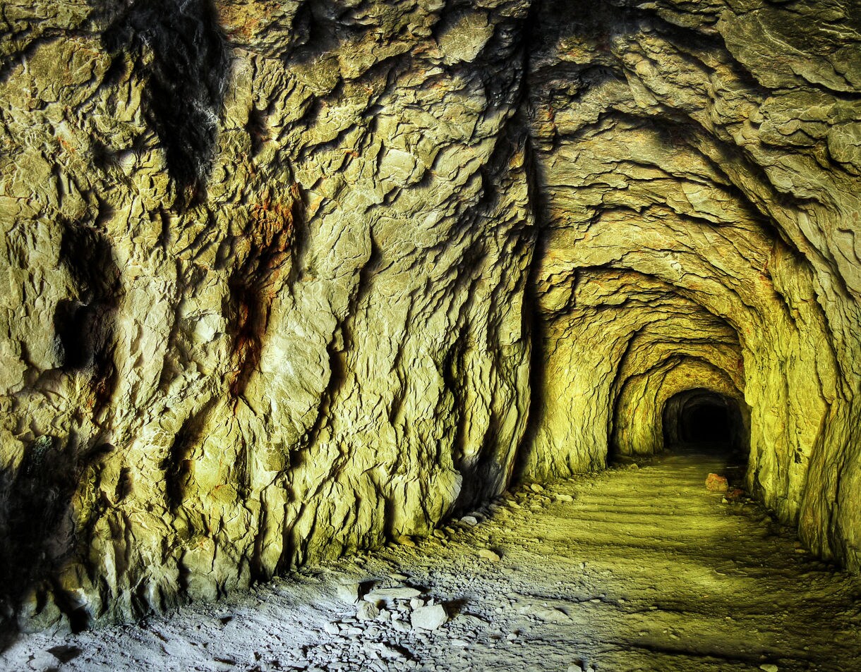 A dimly lit rock tunnel in Rabaul with rough, jagged stone walls and a dirt floor, stretching into darkness and illuminated by warm yellow light.