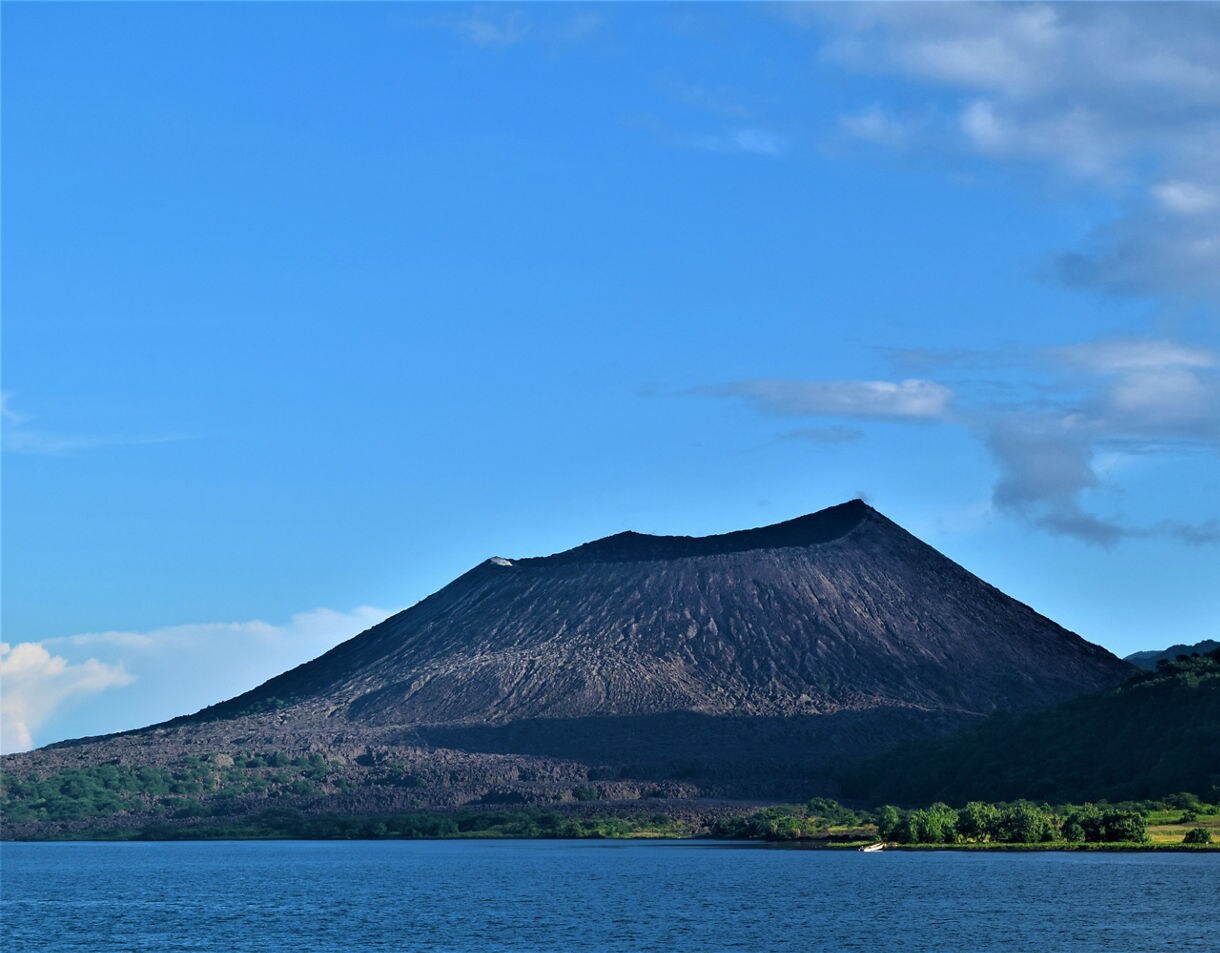 A dark, cone-shaped volcano in Rabaul framed by a bright blue sky, with lush green slopes at its base and a wide expanse of ocean in the foreground.