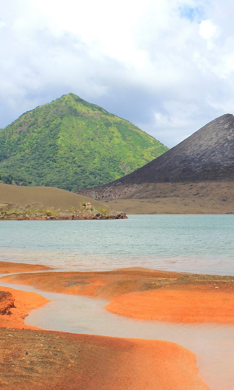 Vertical view of Rabaul’s volcanic landscape showing orange mineral-rich hot springs in the foreground, pale blue water, and two green and dark ash-covered volcanic mountains rising under a cloudy sky.