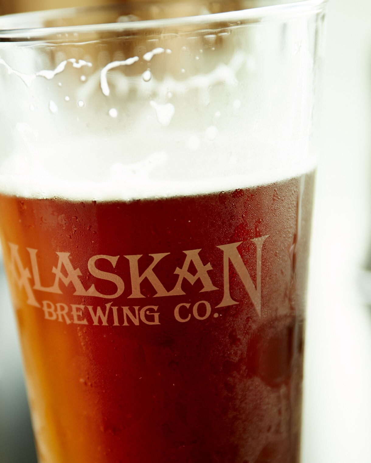 A pint glass filled with amber-colored beer prominently displays the 'Alaskan Brewing Co.' text. The glass is set on a reflective surface, with another beverage blurred in the background. The scene suggests a casual, relaxed setting, possibly in a bar or restaurant. The focus is on the beverage and the clear branding on the glass.
