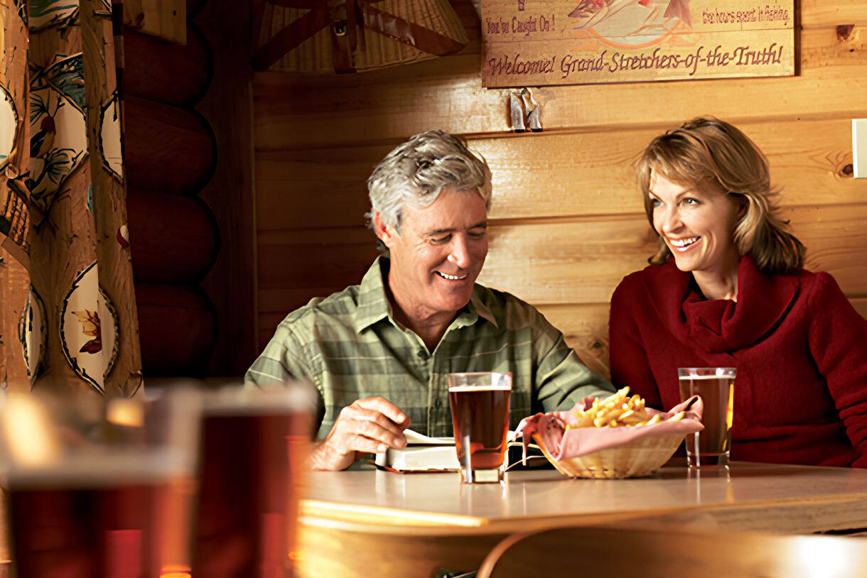 A couple sits together at a wooden table in a cozy, cabin-style setting, sharing drinks and snacks. The background features a sign reading 'Fly Fishers' Club' and vintage fishing gear mounted on wood-paneled walls. Warm lighting and earth tones create a relaxed, inviting mood. Several glasses of amber beer are visible, along with a bowl of snacks. The scene evokes leisure and camaraderie in a lodge environment.