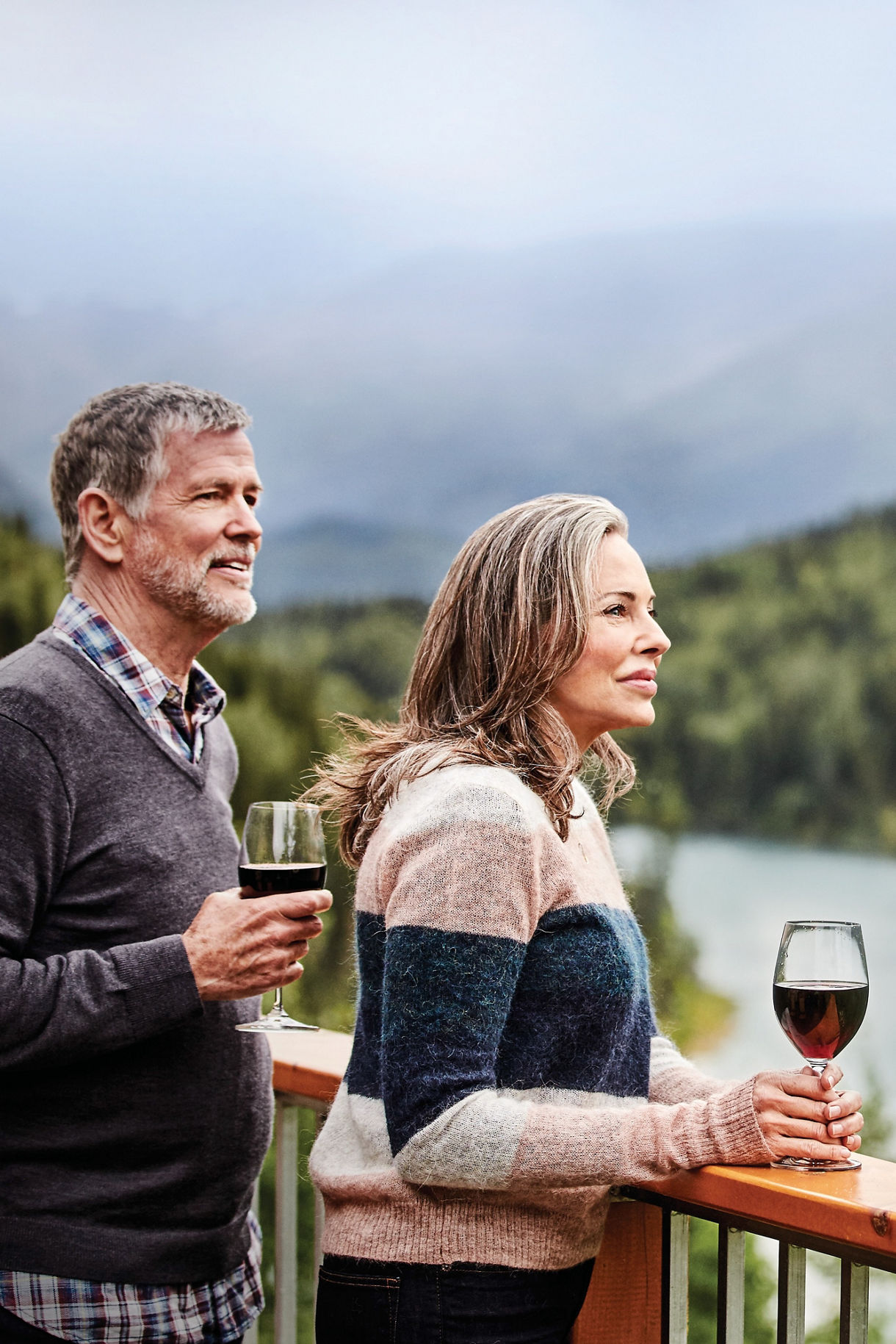 Two adults stand on a wooden balcony overlooking a lush, forested mountain landscape with a lake below. Each person holds a glass of red wine, suggesting a relaxed and leisurely moment. The setting is outdoors, with misty mountains and dense greenery creating a tranquil atmosphere. The overall mood is peaceful and reflective, with soft, natural lighting.