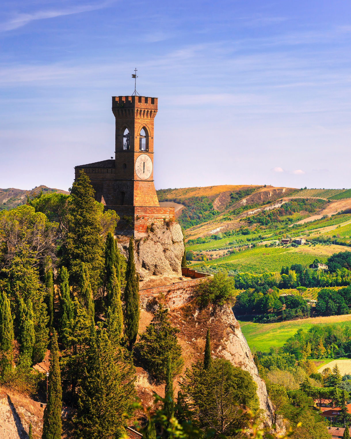 A historic stone clock tower rises dramatically from a rocky hilltop surrounded by tall cypress trees. Below and beyond, rolling green hills and patchwork fields stretch into the distance under a clear blue sky. Small farmhouses and village buildings are scattered across the landscape, adding warmth and human presence to the rural scene. The bright daylight and vivid colors create a serene, picturesque view of the Mediterranean countryside.