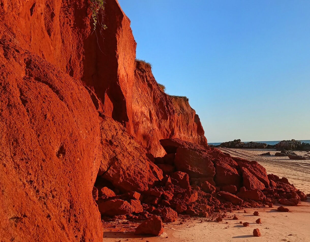 A sandy beach scattered with small stones sits below tall layered cliffs, with calm blue water and a bright sky overhead.
