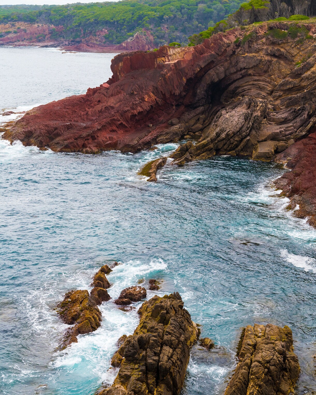 A coastal scene featuring layered red and brown rock formations curving into a cave-like fold, bordered by turquoise waves and surrounded by green, forested headlands.