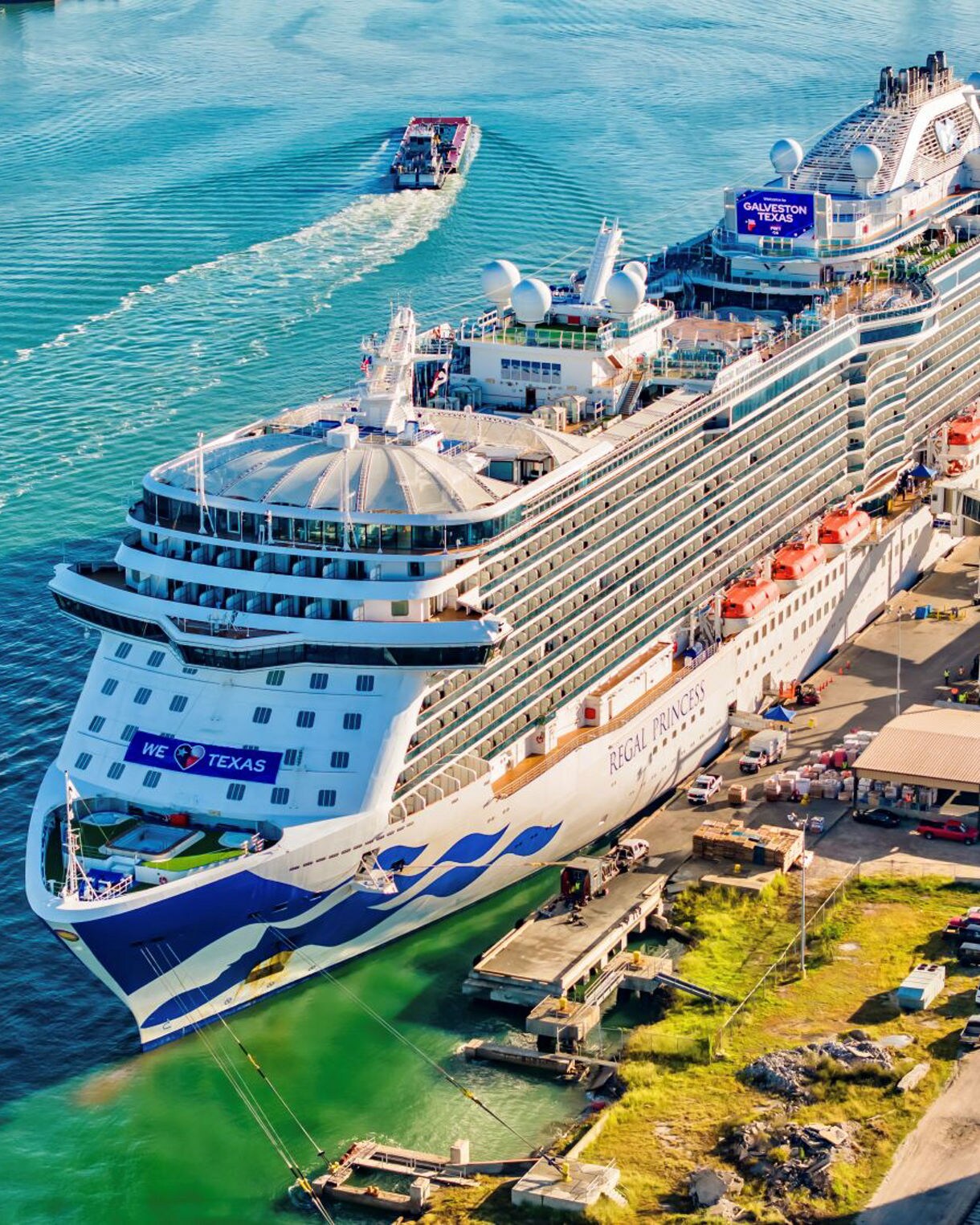 Aerial view of Regal Princess cruise ship docked at Galveston port, with a smaller boat approaching from the left, showcasing the ship's white and blue design and prominent "WE HEART TEXAS" banner. 