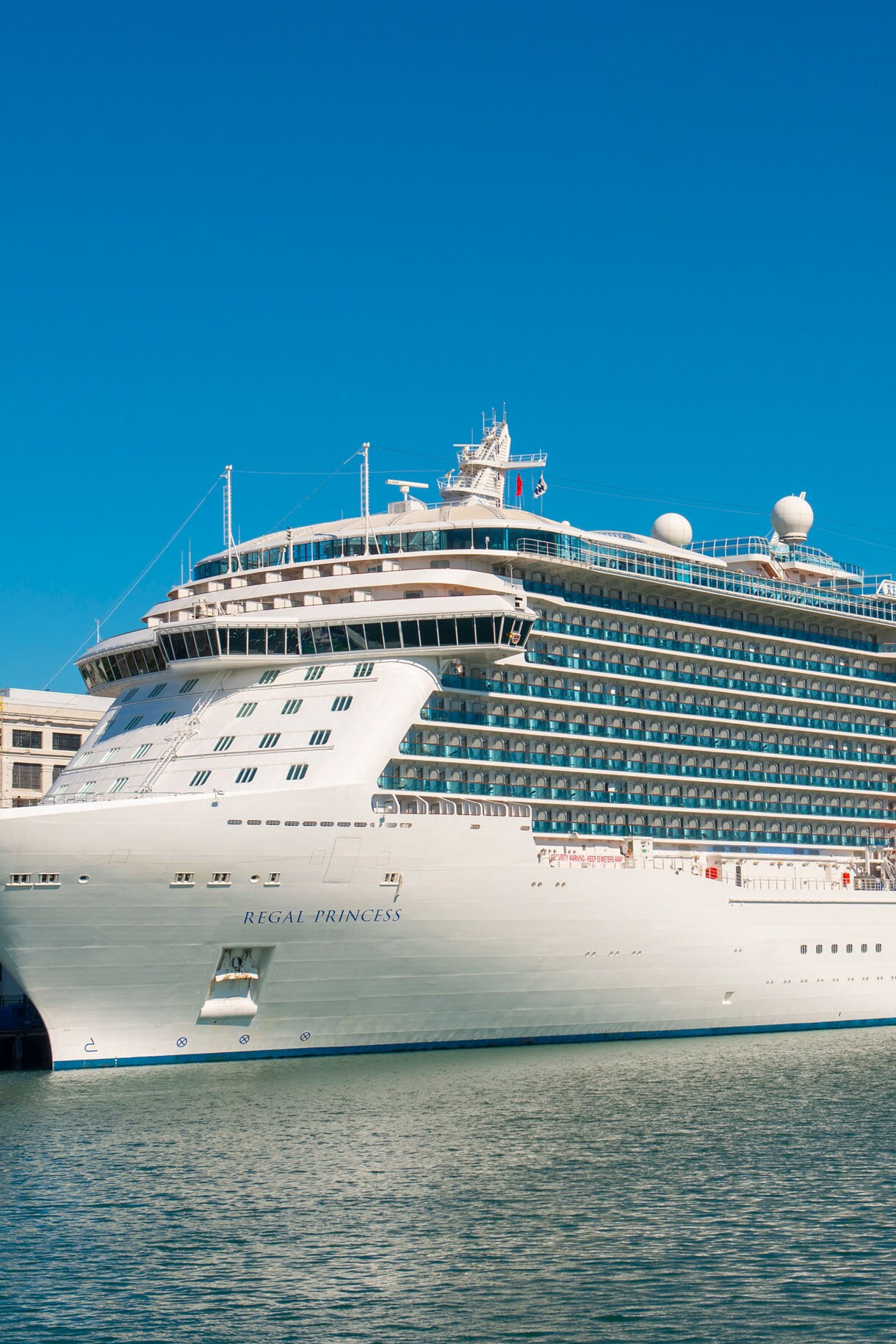 The Regal Princess cruise ship docked at port, with its sleek white exterior and rows of balconies gleaming under a clear blue sky.