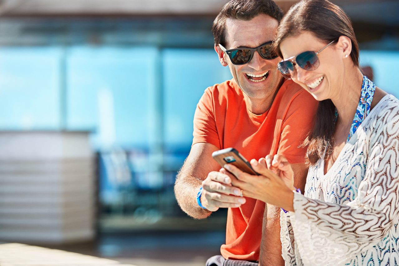 A man and woman wearing sunglasses smile while looking together at a smartphone on the ship's deck.