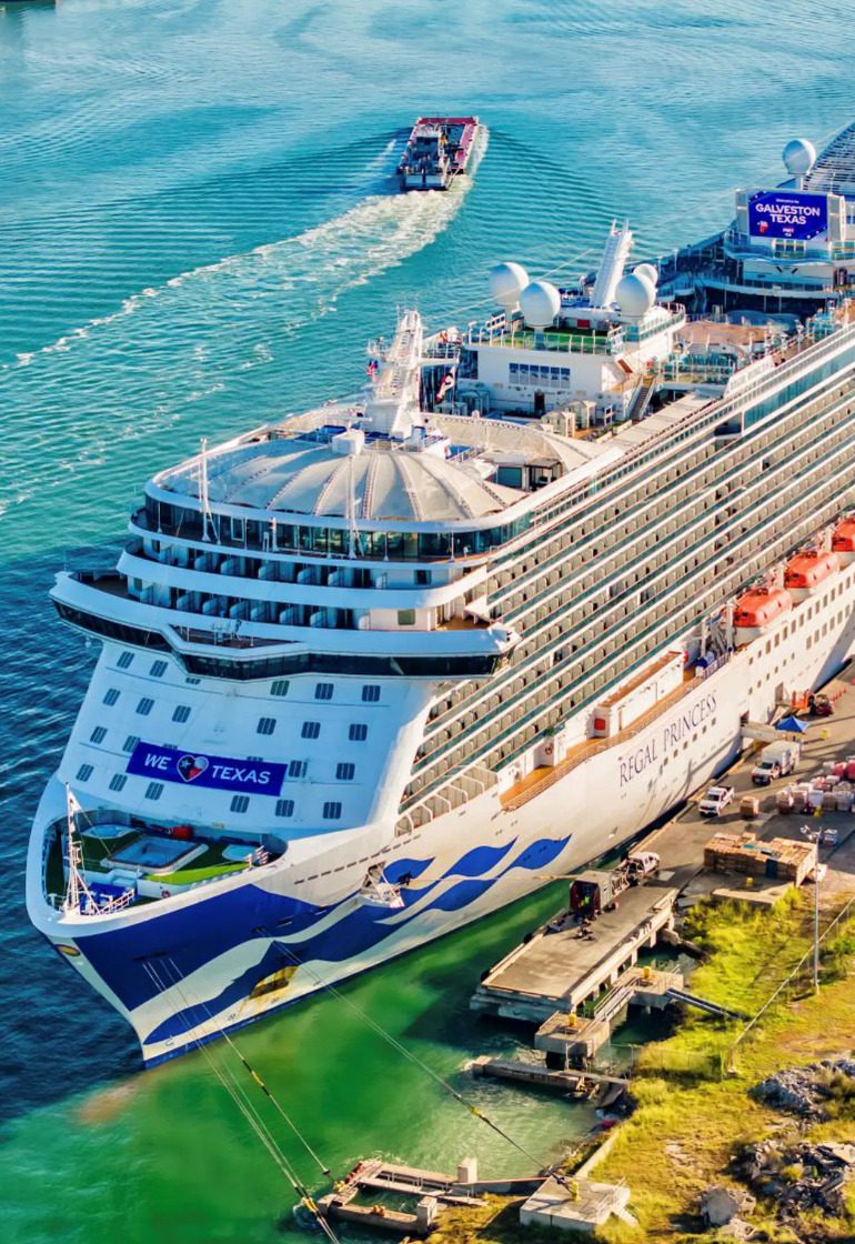 Aerial view of the Regal Princess cruise ship docked in Galveston, Texas, with a vibrant blue and white hull, rows of balconies, and a welcoming message reading "We ♥ Texas." A smaller boat travels nearby, leaving a trail in the water.