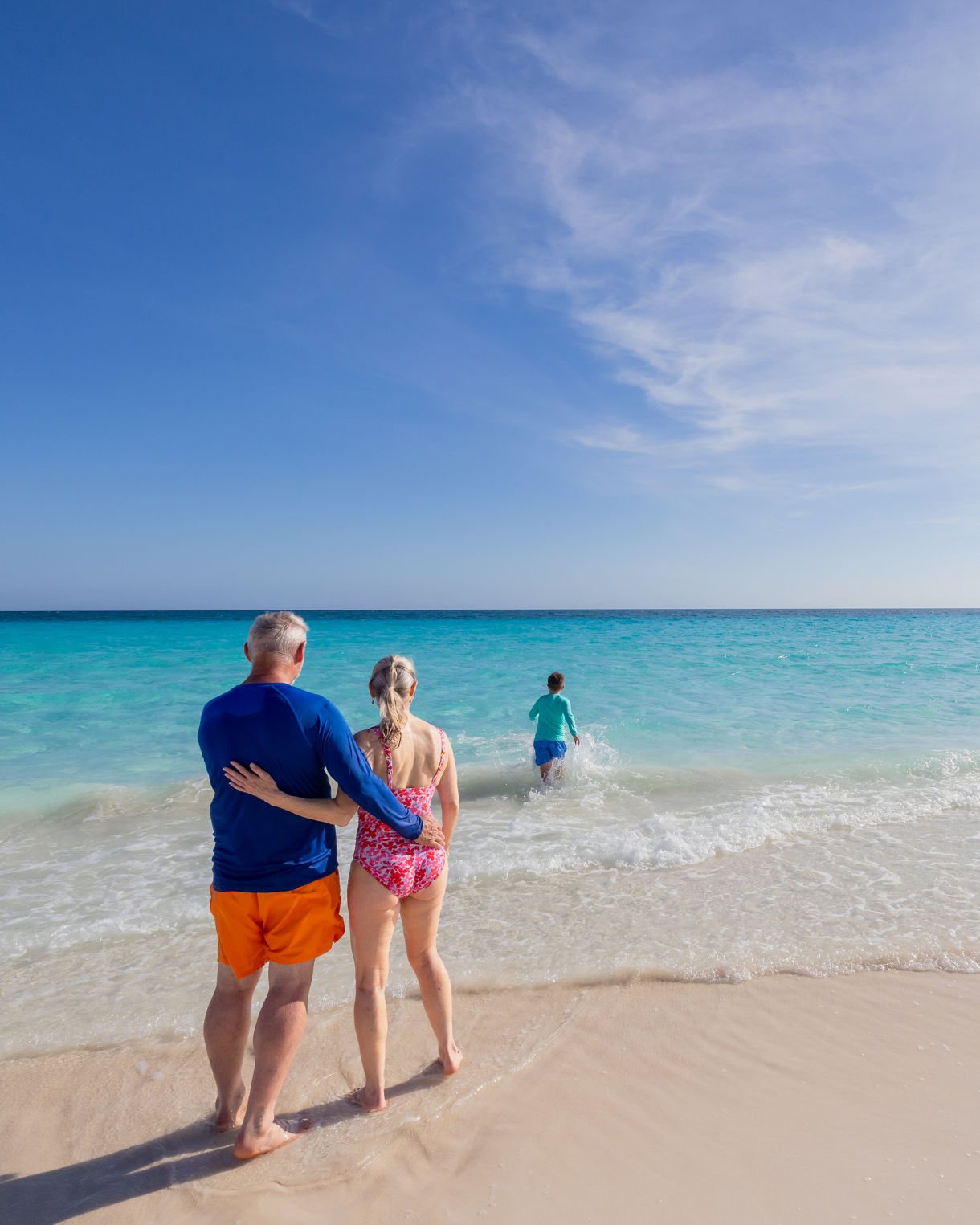 An older couple stands together on a sandy beach, looking out at the turquoise ocean.
