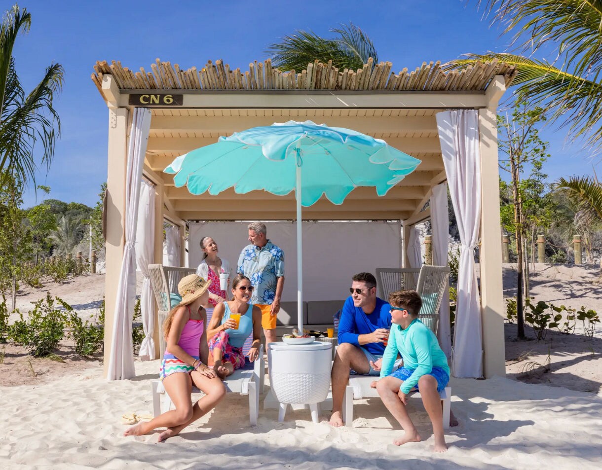 A multigenerational family relaxes together under a shaded cabana on a sunny beach
