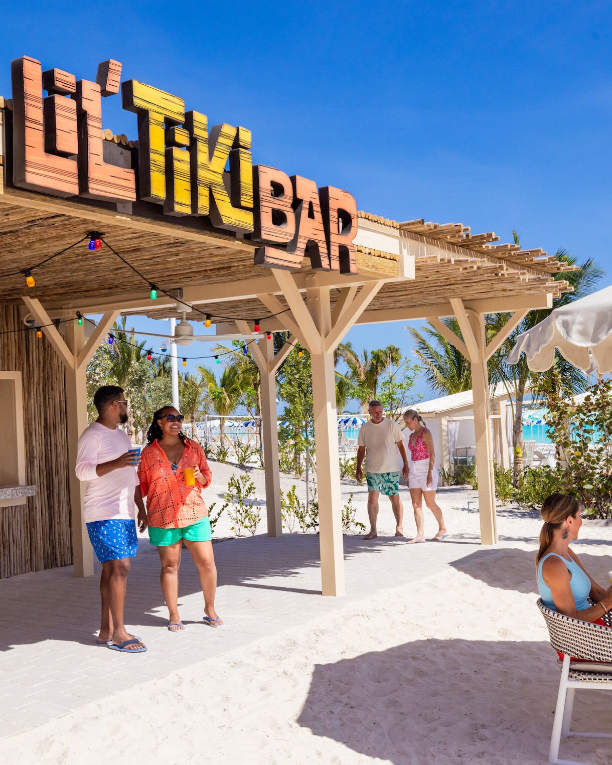 A group of adults socialize at an open-air tiki bar on a sunny beach.