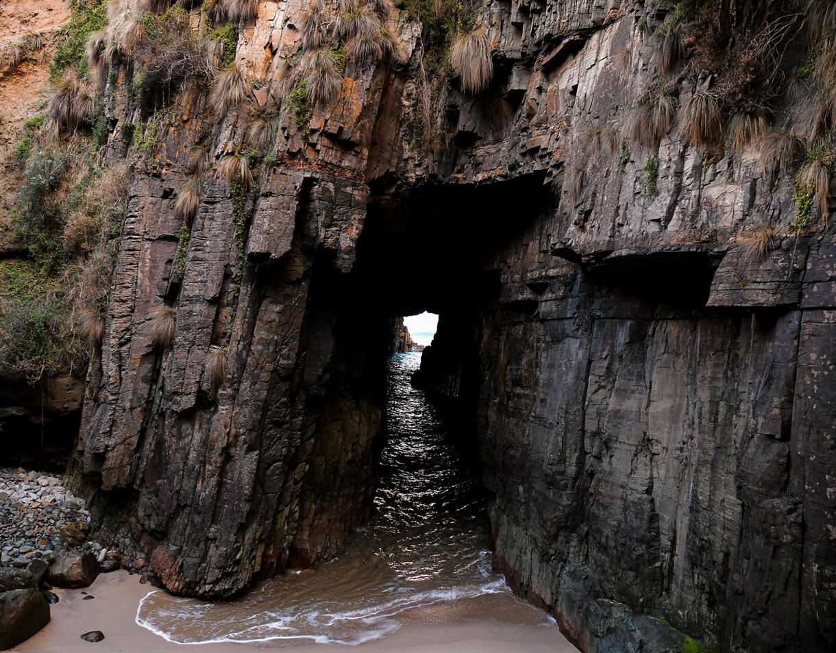 A narrow sea cave carved into tall layered cliffs, with seawater flowing through a dark tunnel toward a small opening of light.