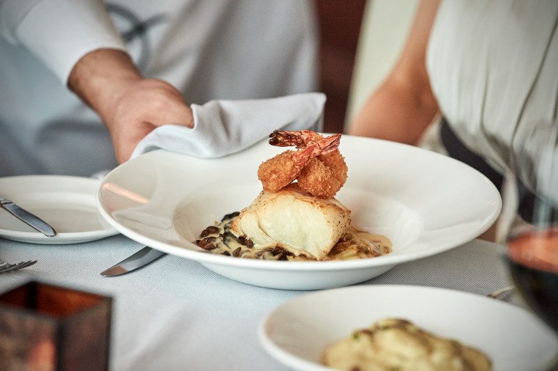 An elegant plated dish shows a white fish fillet topped with golden-brown breaded shrimp, served on creamy mushroom sauce in a white plate, with a server's hand visible.