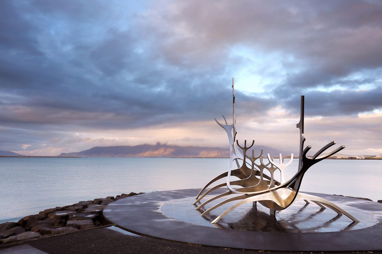 Metal sculpture on a platform in a fjord, surrounded by calm water and a distant mountain range under a cloudy sky.