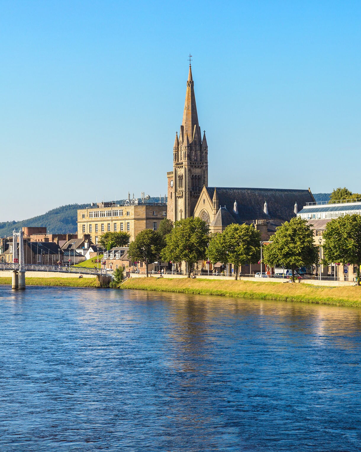 View of the River Ness in Inverness, Scotland, with tree-lined banks, church spires and hills rising in the distance under a clear blue sky.