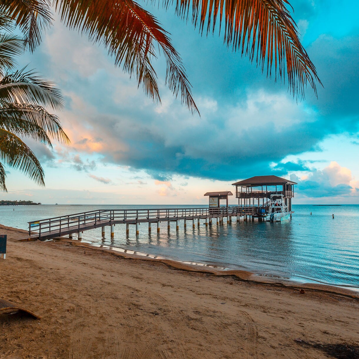 Wooden pier extending into calm water at sunset with palm fronds overhead and covered pavilion structure