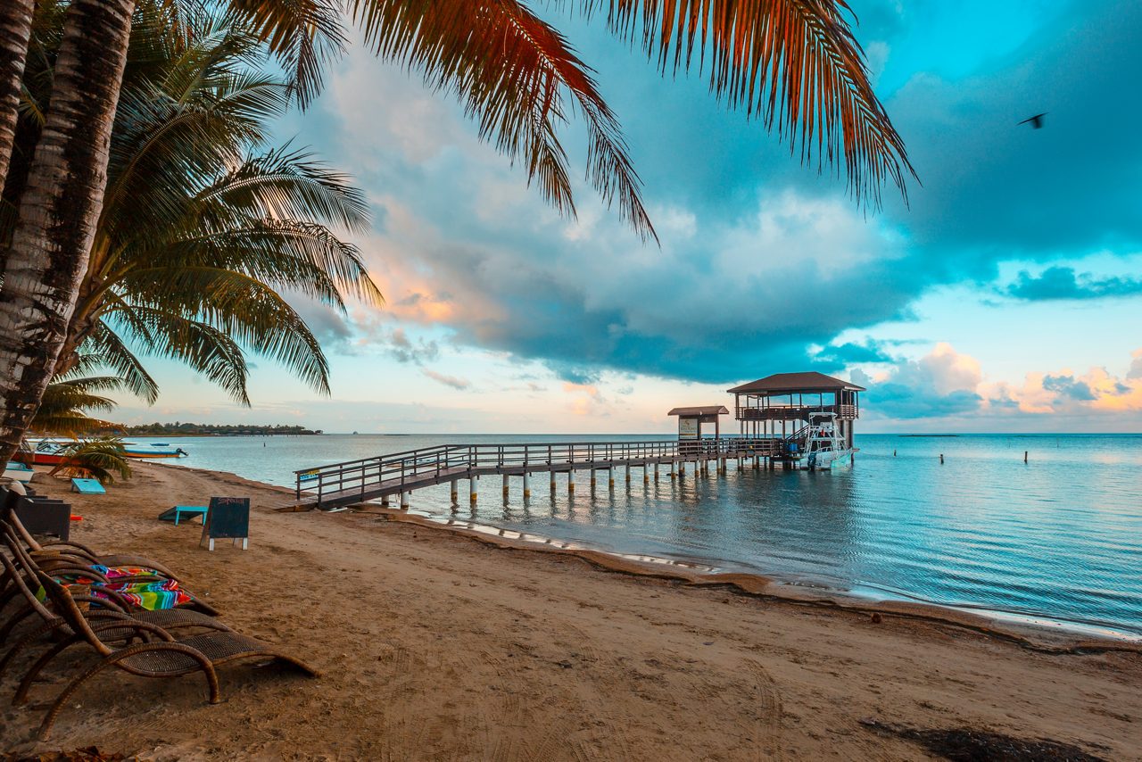 A tropical beach in Roatán, Honduras at sunset, featuring a wooden pier extending over calm waters.