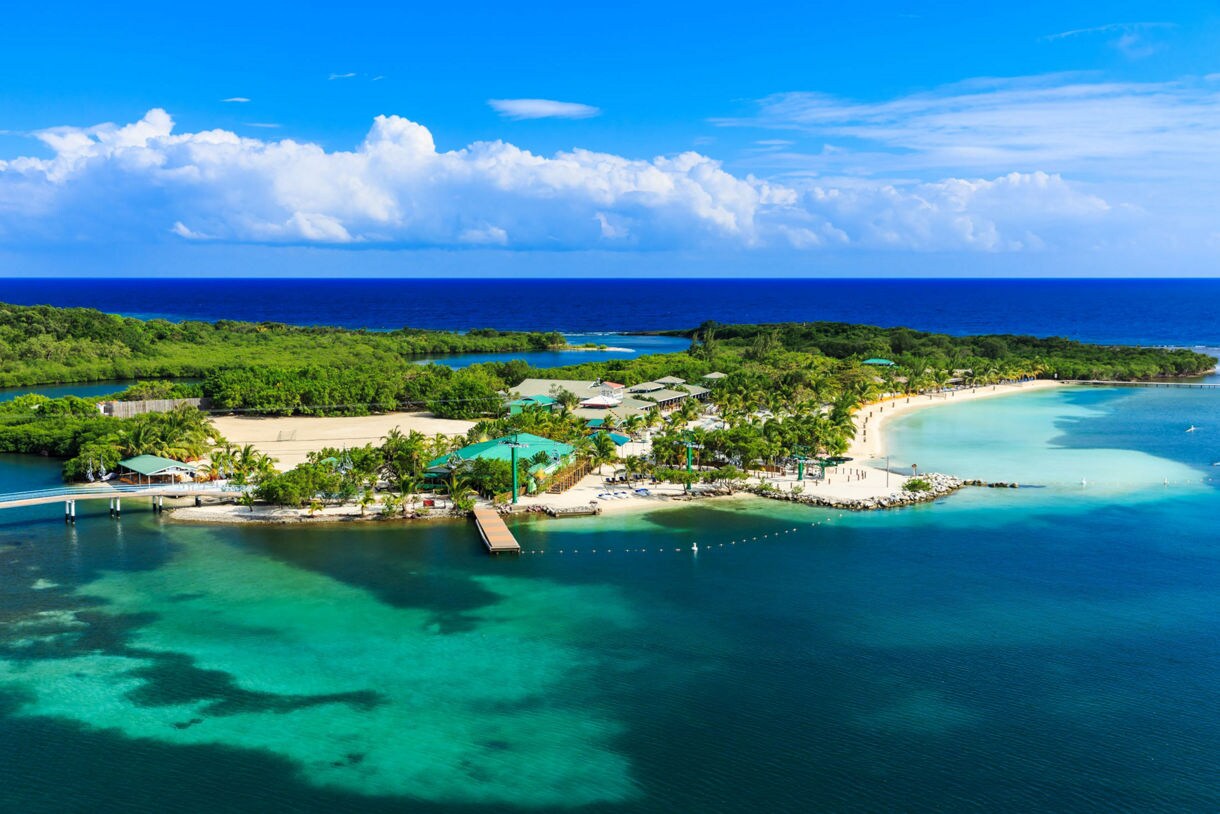 A scenic view of Oak Ridge, Roatán, featuring brightly colored stilt houses along a calm waterway, with a small boat navigating through and the ocean stretching to the horizon under a golden sunset sky.