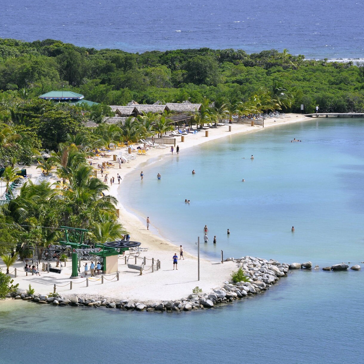 Aerial view of Mahogany Bay, Roatán, featuring the beach, tropical palms and an aerial chairlift moving through the lush landscape, with visitors enjoying the water and scenic views.