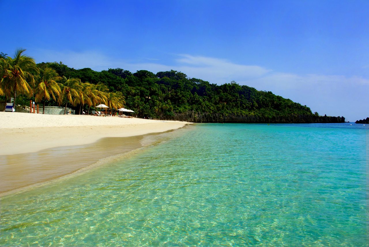 A pristine stretch of West Bay Beach in Roatán, Honduras, featuring crystal-clear turquoise water, soft white sand and a row of palm trees with shaded lounge areas under a bright blue sky.