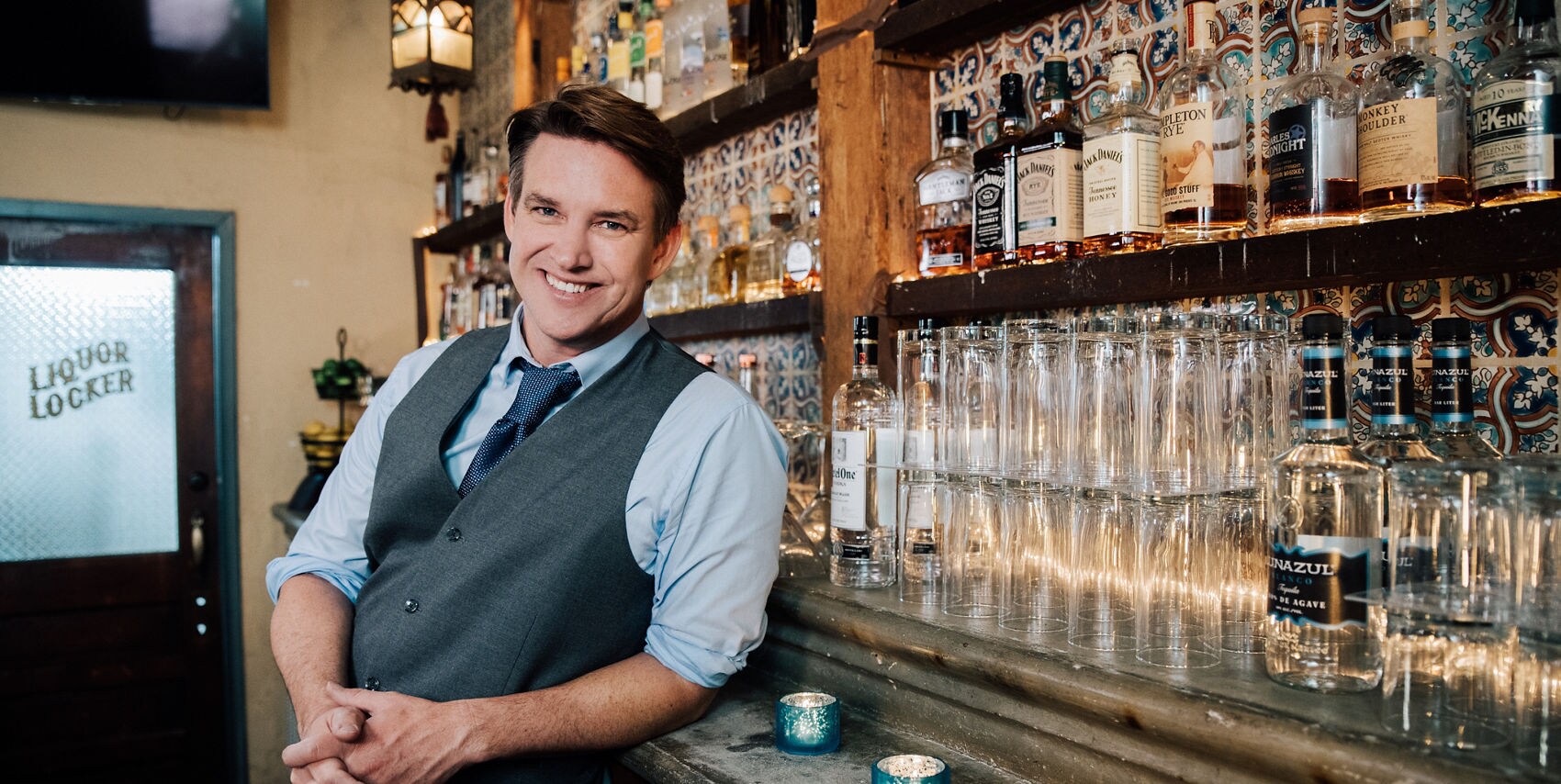 A smiling man in a vest stands confidently in front of a well-stocked bar.