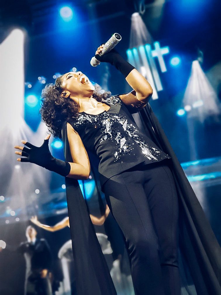Female vocalist in a sparkly black outfit singing passionately into a microphone under blue stage lights