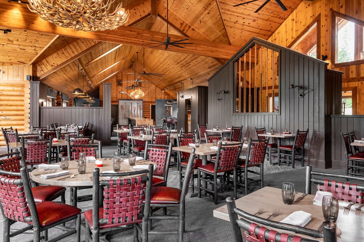 Spacious restaurant dining area features wooden walls and ceiling with exposed beams, creating a cozy, rustic atmosphere. Tables are set with glassware, napkins, and cutlery, while red woven chairs add a pop of color. Large windows allow natural light to fill the space, and antler-style chandeliers enhance the lodge-like ambiance. No visible text or numbers are present in the image.