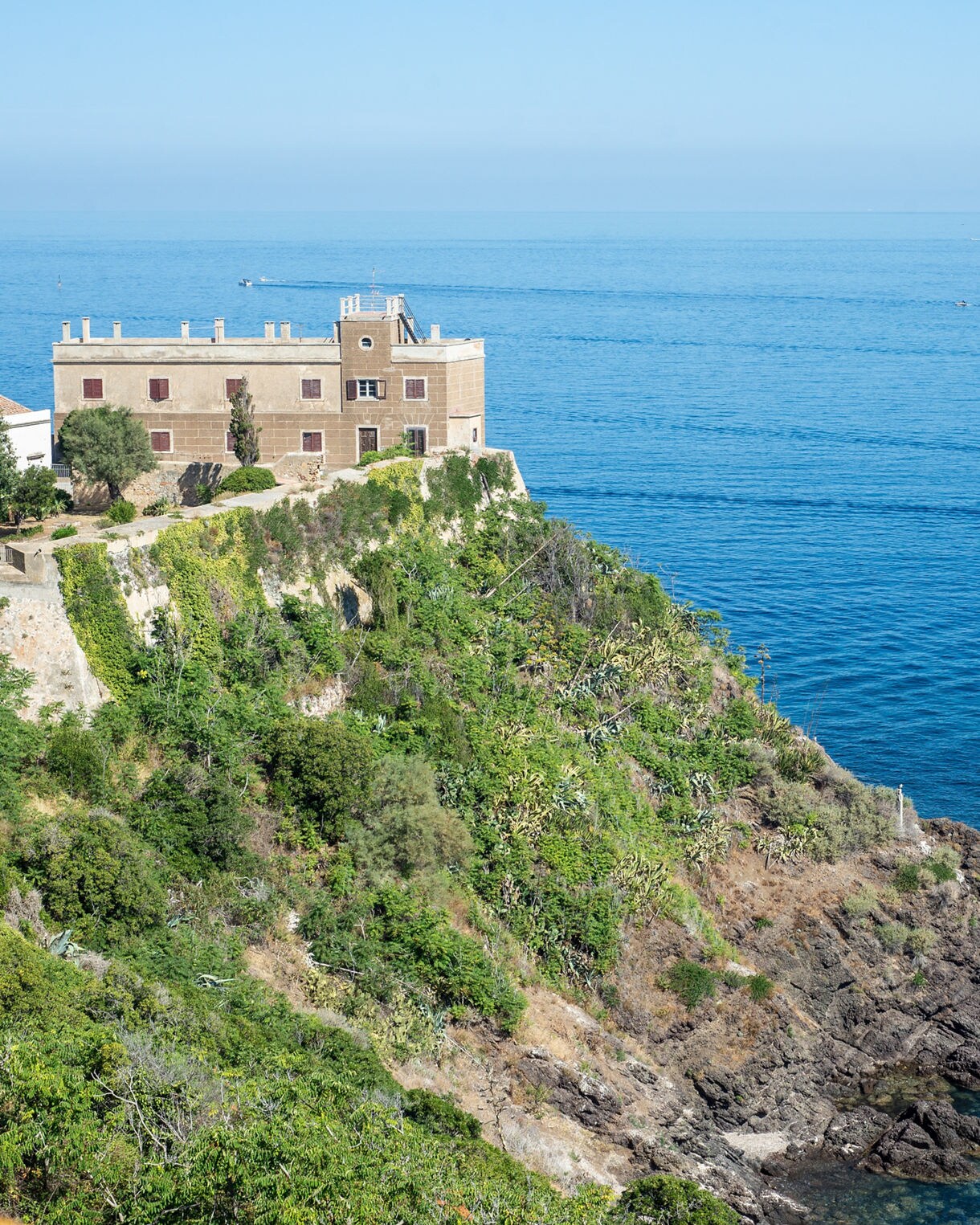 Historic stone villa perched atop a lush cliff on Elba Island, overlooking calm blue Mediterranean waters with rocky shoreline below.