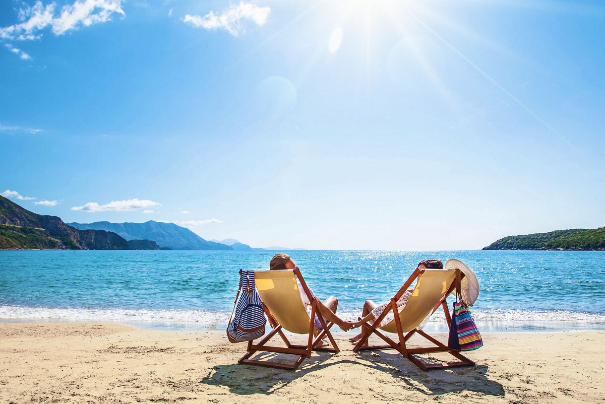 Couple lounging in beach chairs on a sandy shore, holding hands and facing a calm blue sea with green hills in the distance, under a bright sunny sky.