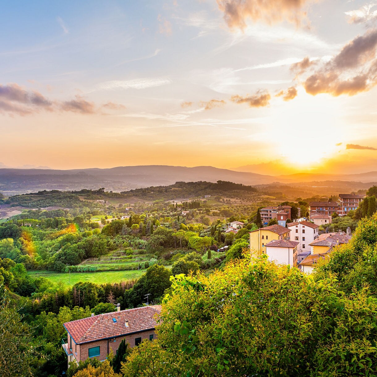 Rolling countryside with fields, trees, and small houses at sunset in Tuscany, Italy.