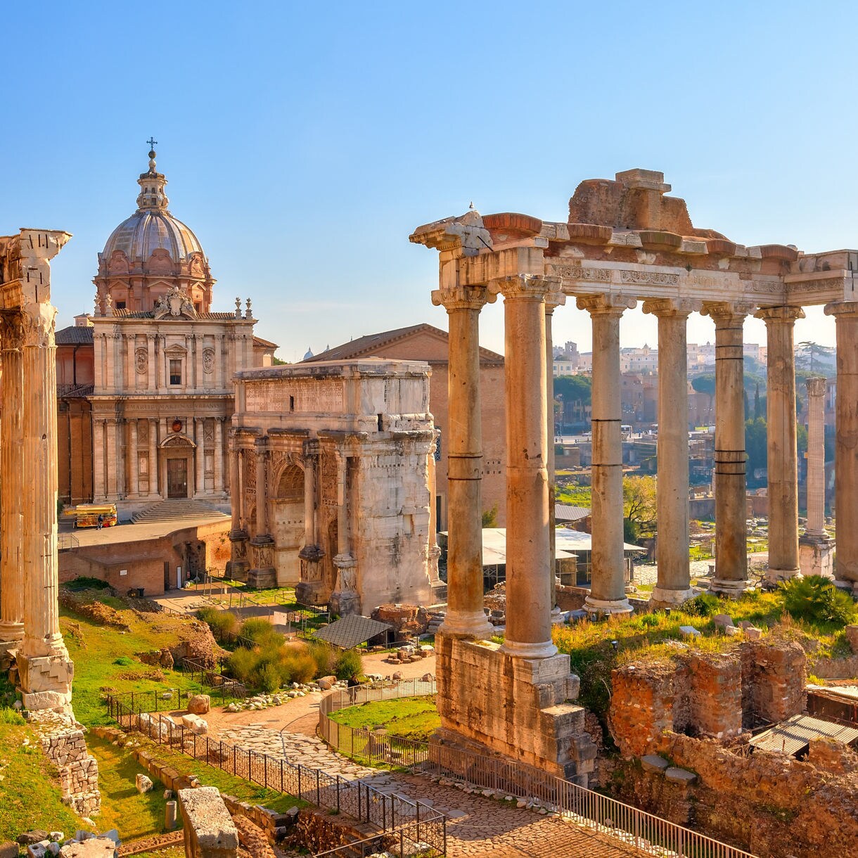 Ancient ruins of the Roman Forum with tall columns, historic buildings, and stone pathways under a clear sky.